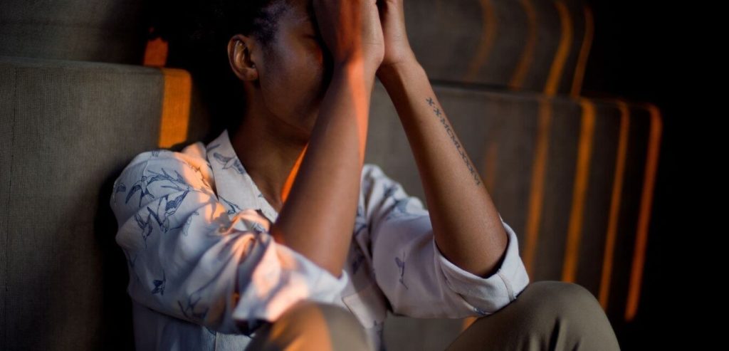 A brown skinned man sits on the floor with his hands on his forehead